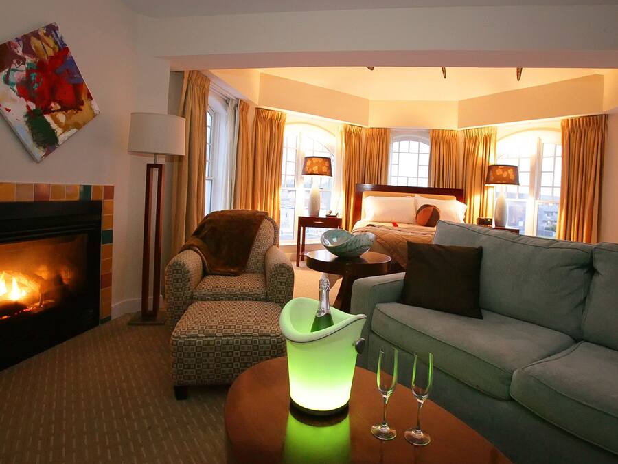 Una habitación de hotel con chimenea, un cubito de hielo verde, dos vasos, un sofá, un sillón estampado, una mesita de noche con una lámpara y un cuadro en la pared.
