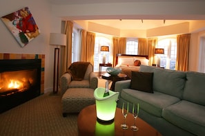 A hotel room with a fireplace, a green ice bucket, two glasses, a sofa, a patterned armchair, a side table with a lamp, and a painting on the wall.