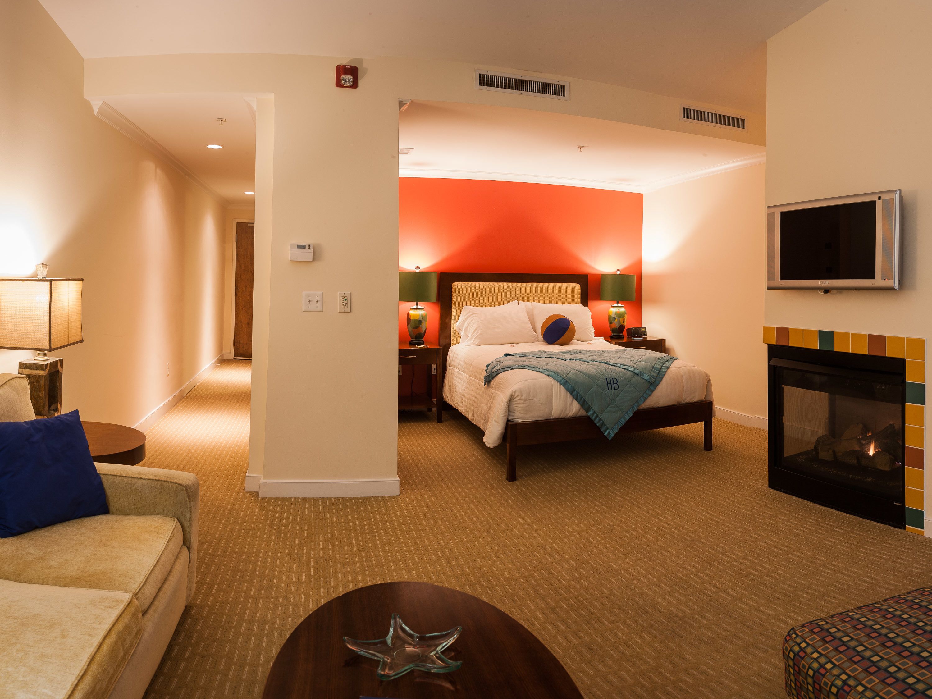 A hotel room with a bed, a TV, a fireplace, and a hallway leading to another room.