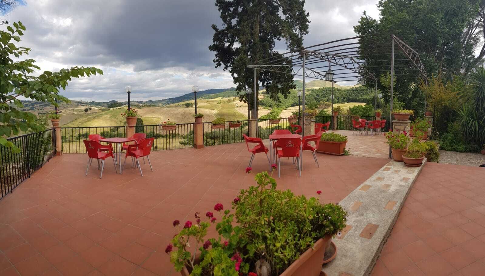 Um pátio externo com cadeiras e mesas vermelhas, rodeado por vasos de plantas e uma pérgola de metal.
