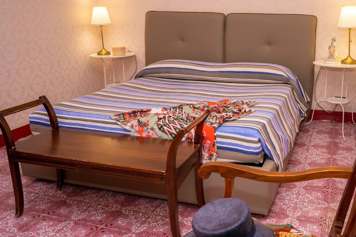 A hotel room with a double bed, a wooden nightstand, a chair, and a small table with a lamp.