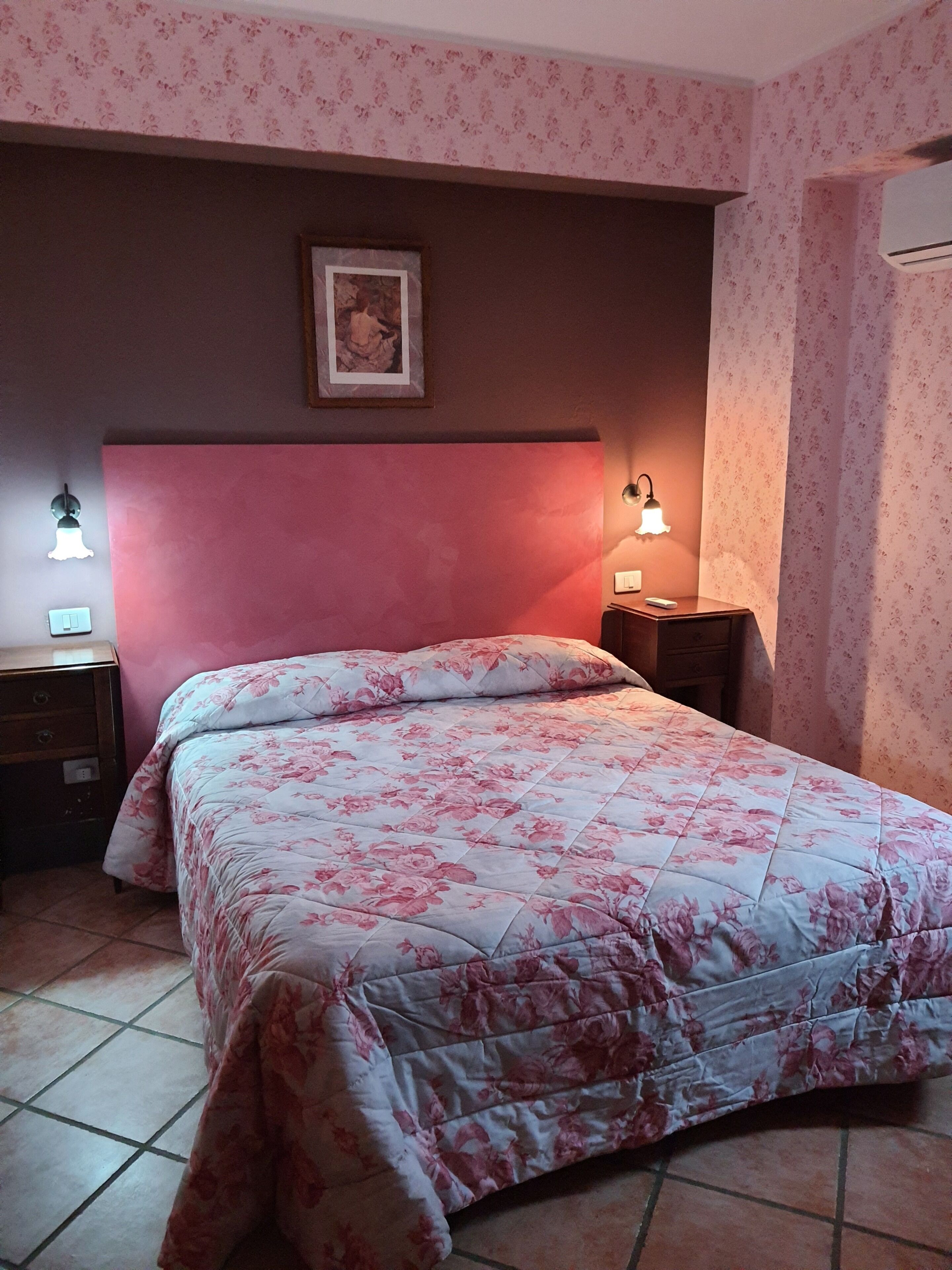 Un dormitorio con una cama, dos mesitas de noche, una mesita auxiliar, un cuadro en la pared y un equipo de aire acondicionado.
