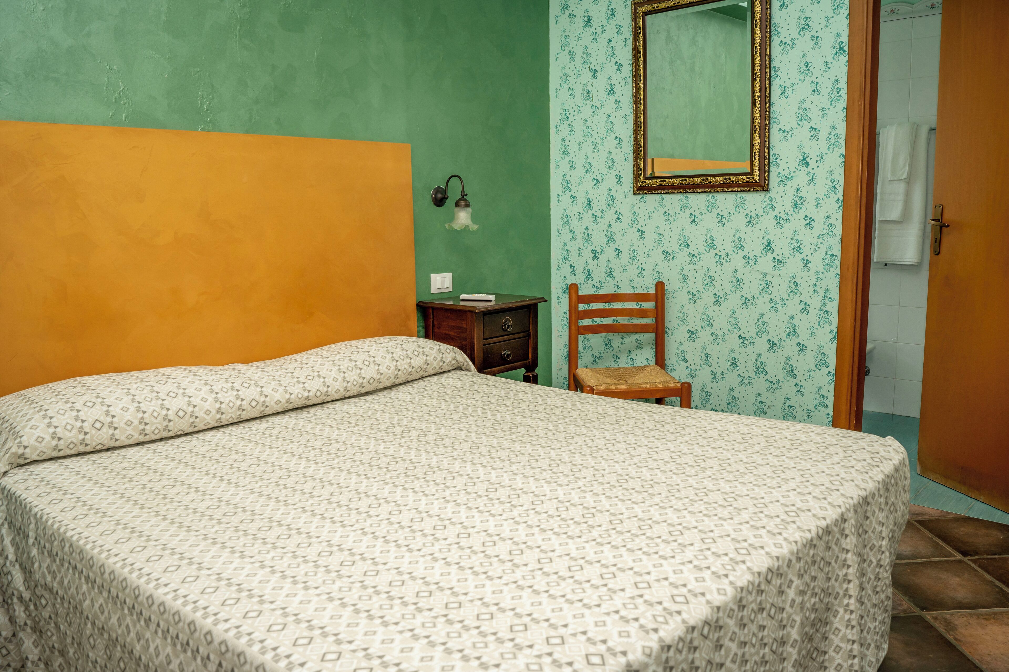 A room with a bed, a nightstand, a chair, a mirror, and a door leading to a bathroom.