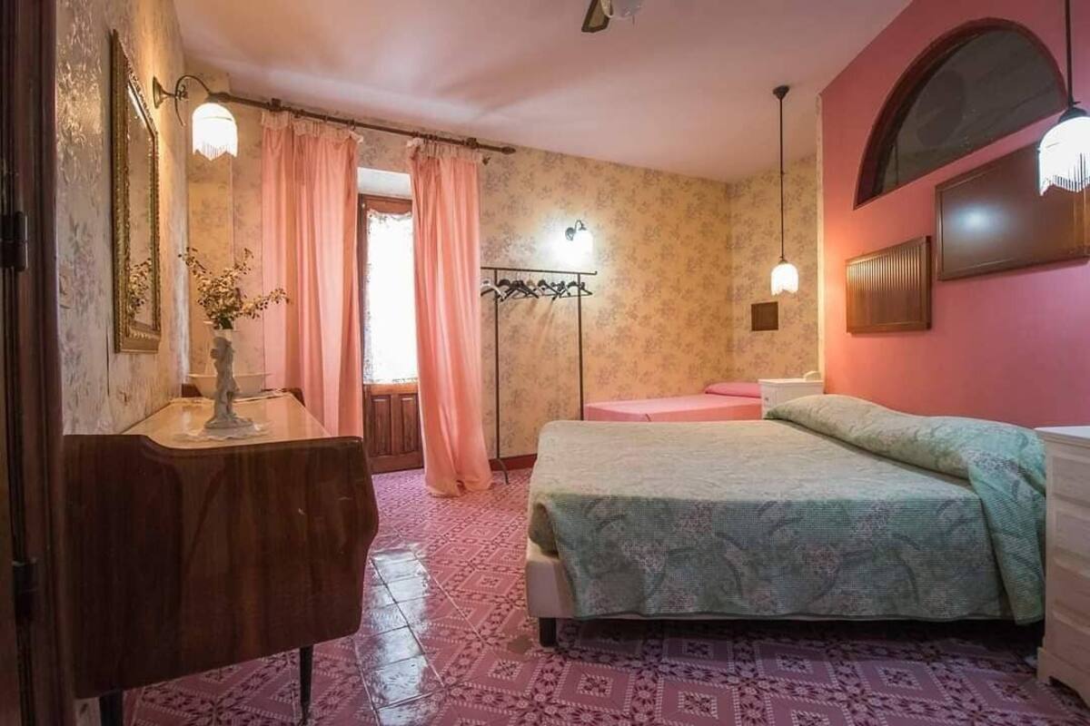 A bedroom with a bed, a vanity with a mirror, a wall-mounted light, and a window with curtains.