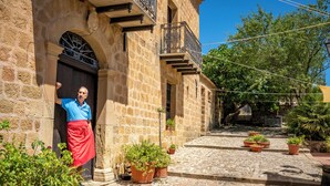 Edifício de pedra com um homem de camisa azul e avental vermelho parado na entrada. Há plantas em vasos no chão de calçada.