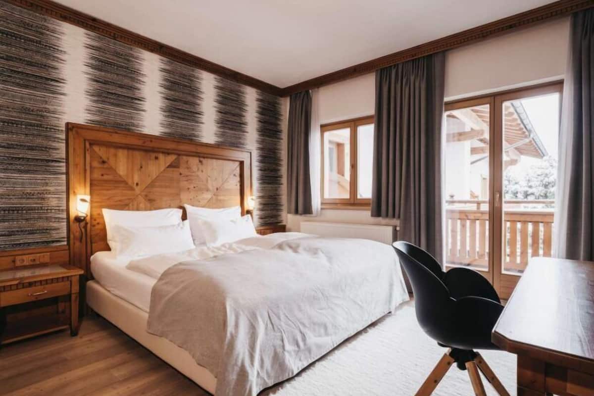 Une chambre à coucher avec une tête de lit en bois, un lit recouvert de draps blancs, une table de chevet, une table en bois et une vue sur un balcon par la fenêtre.
