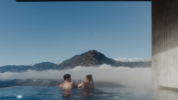 Ein Paar in einer heißen Quelle, im Hintergrund ein Berg mit verschneiten Gipfeln.