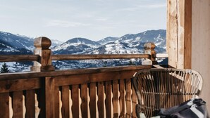 Ein Holzbalkon mit Blick auf schneebedeckte Berge. Zwei Korbstühle und ein kleiner Tisch mit Blumen stehen bereit.