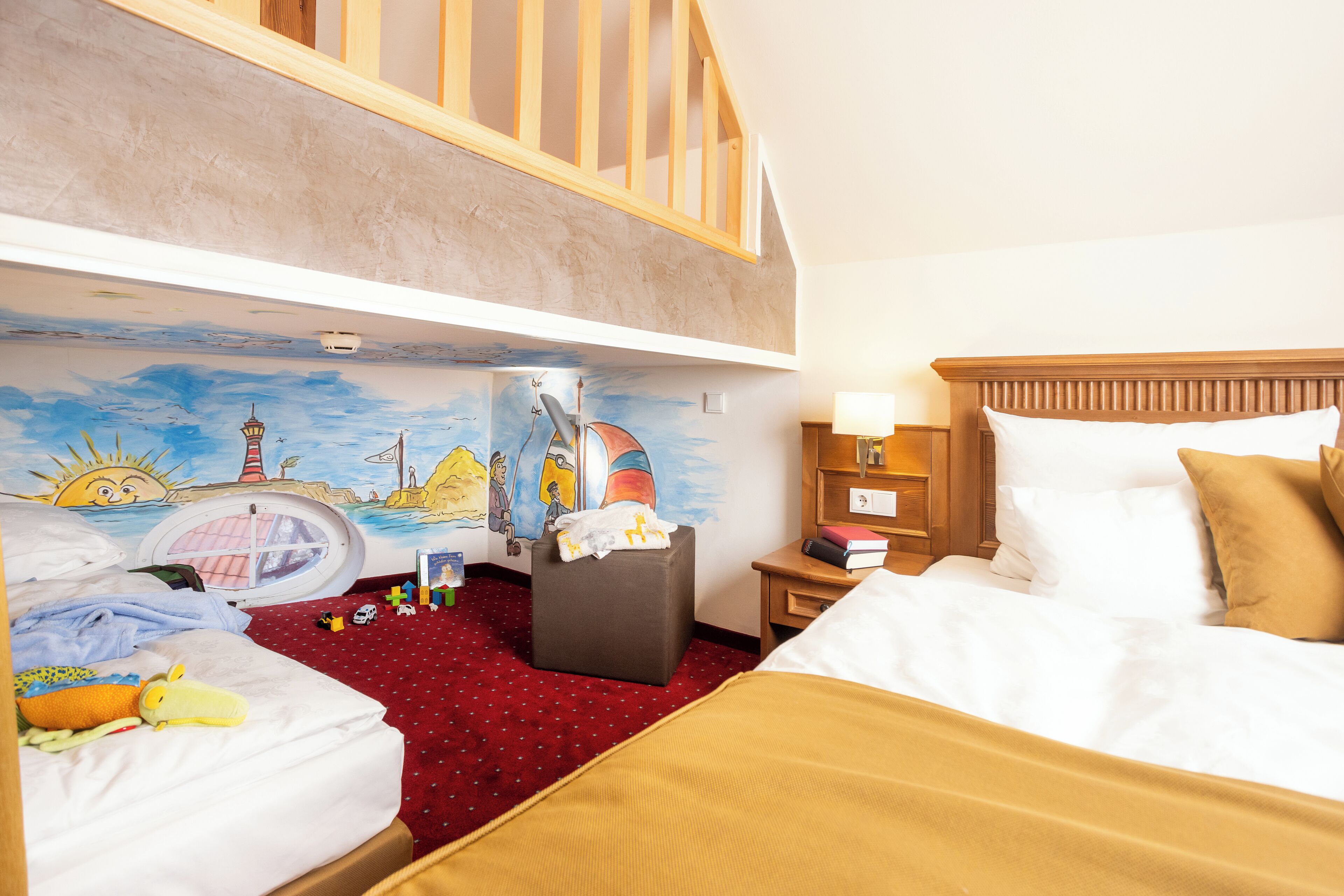 A hotel room with a loft bed, a small desk, a nightstand, a window, and a mural on the wall.