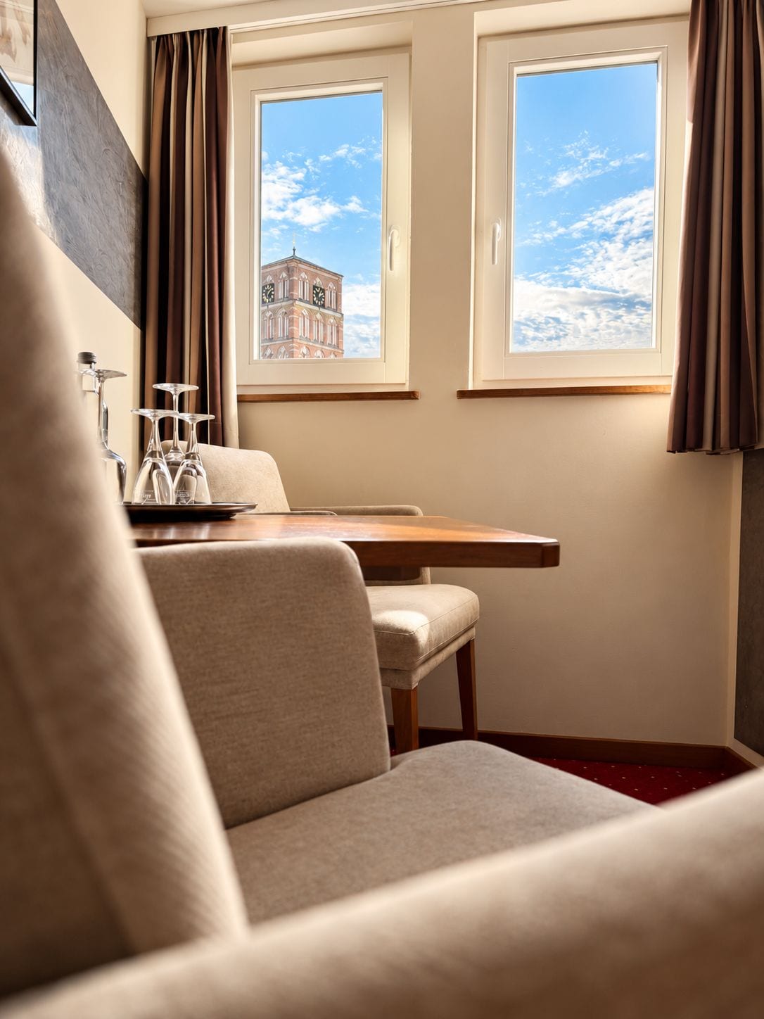 A hotel room with a bed, a table, and two windows with a view of a clock tower.