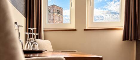 A hotel room with a bed, a table, and two windows with a view of a clock tower.