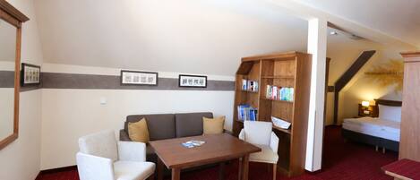 Una habitación con sofá, dos sillones, una mesa, estantería, cama y alfombra roja.