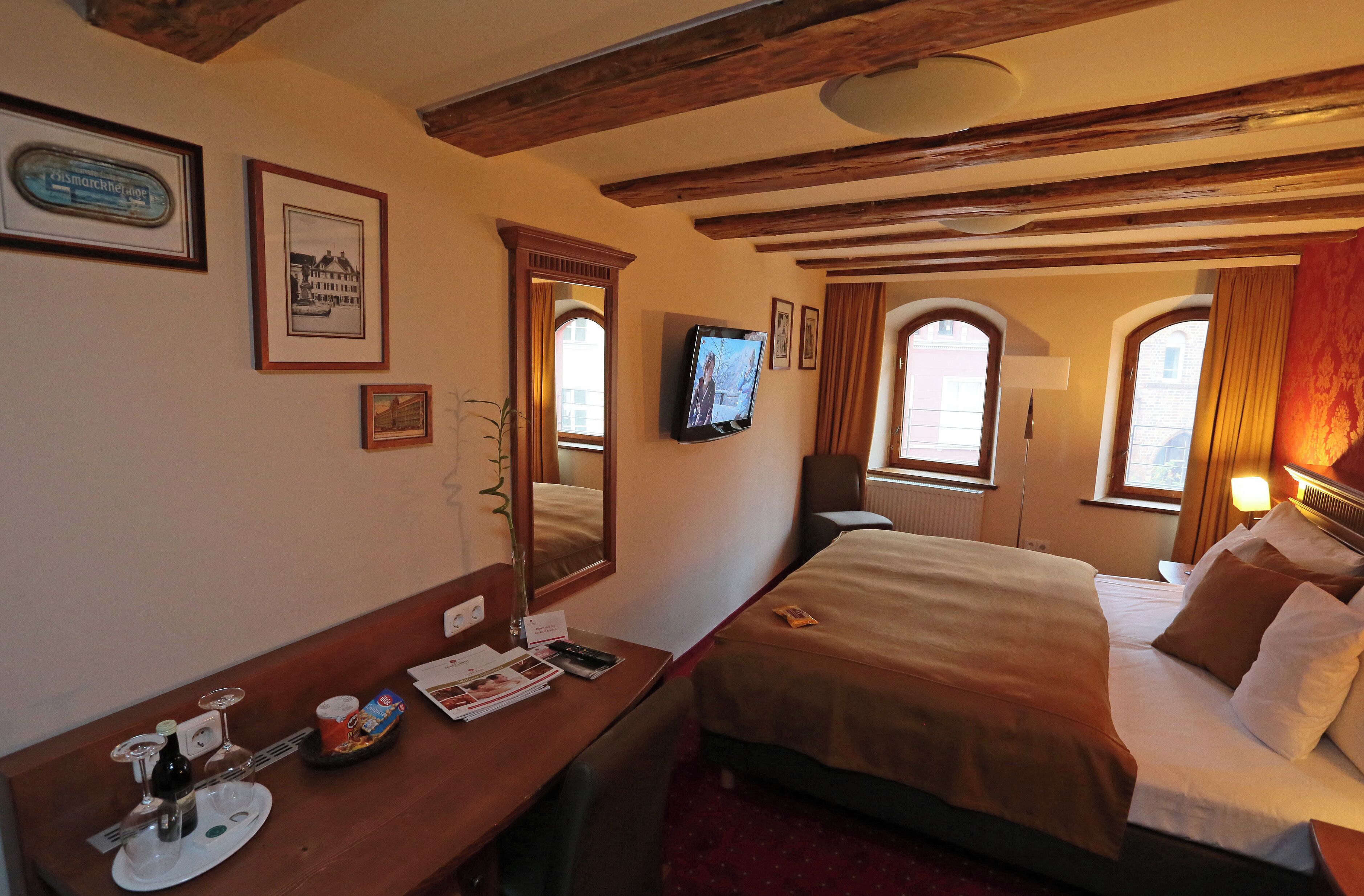 Habitación de hotel con techo de madera, cama con cubrecama marrón, escritorio con un plato de comida y bebidas, televisor y cuadros en la pared.