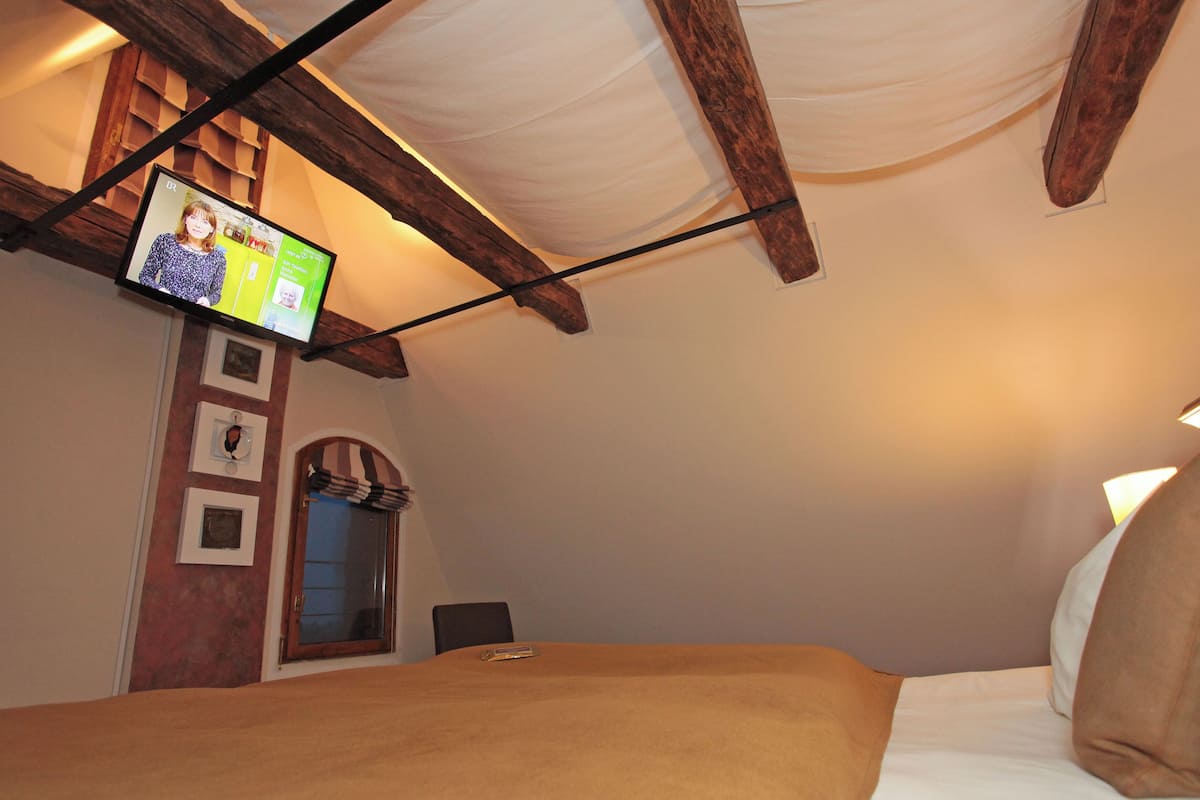 A bedroom with a bed, a television mounted on the wall, wooden beams, and a window with a curtain.