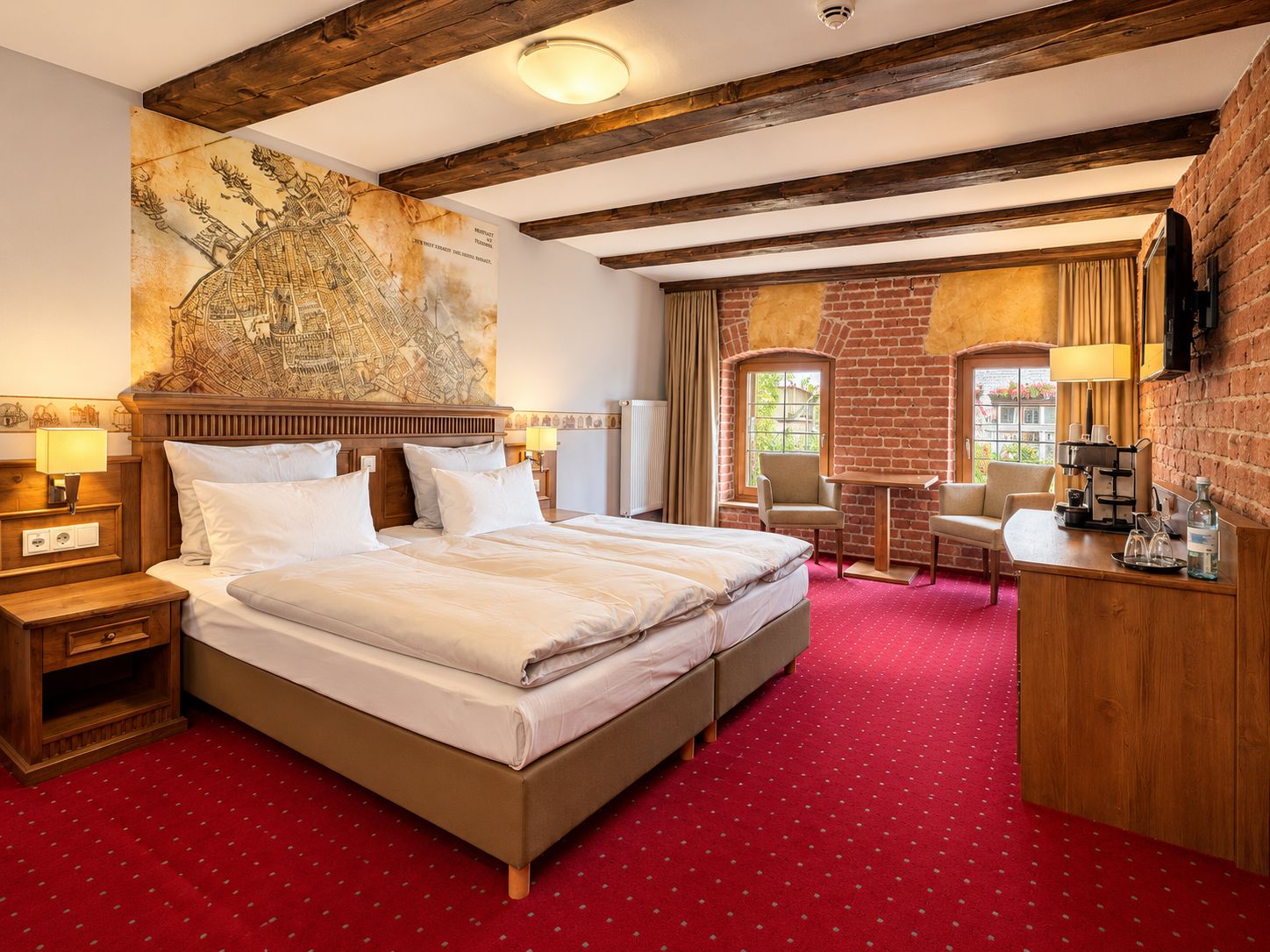 A hotel room with a large bed, a wooden headboard, a red carpet, a map mural, a brick wall, and a window with curtains.