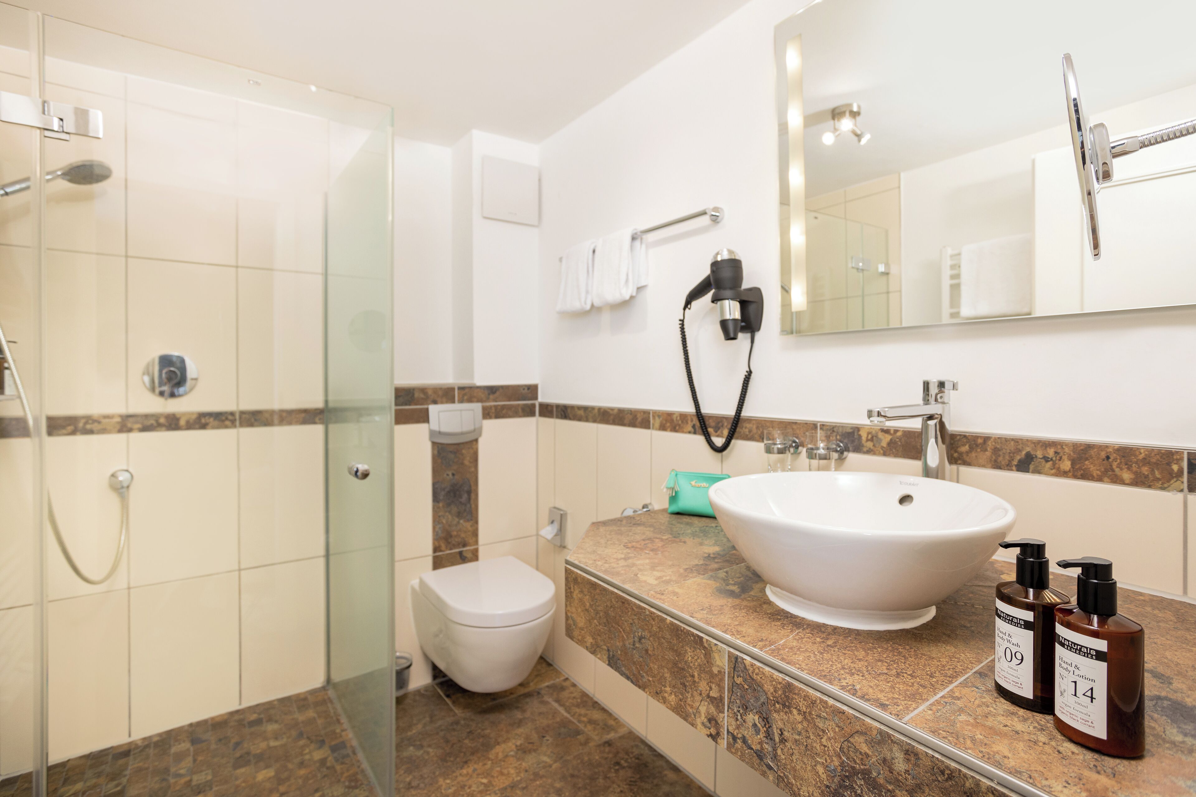 A bathroom with a glass shower enclosure, a white toilet, a large white sink, and two bottles of soap on the countertop.