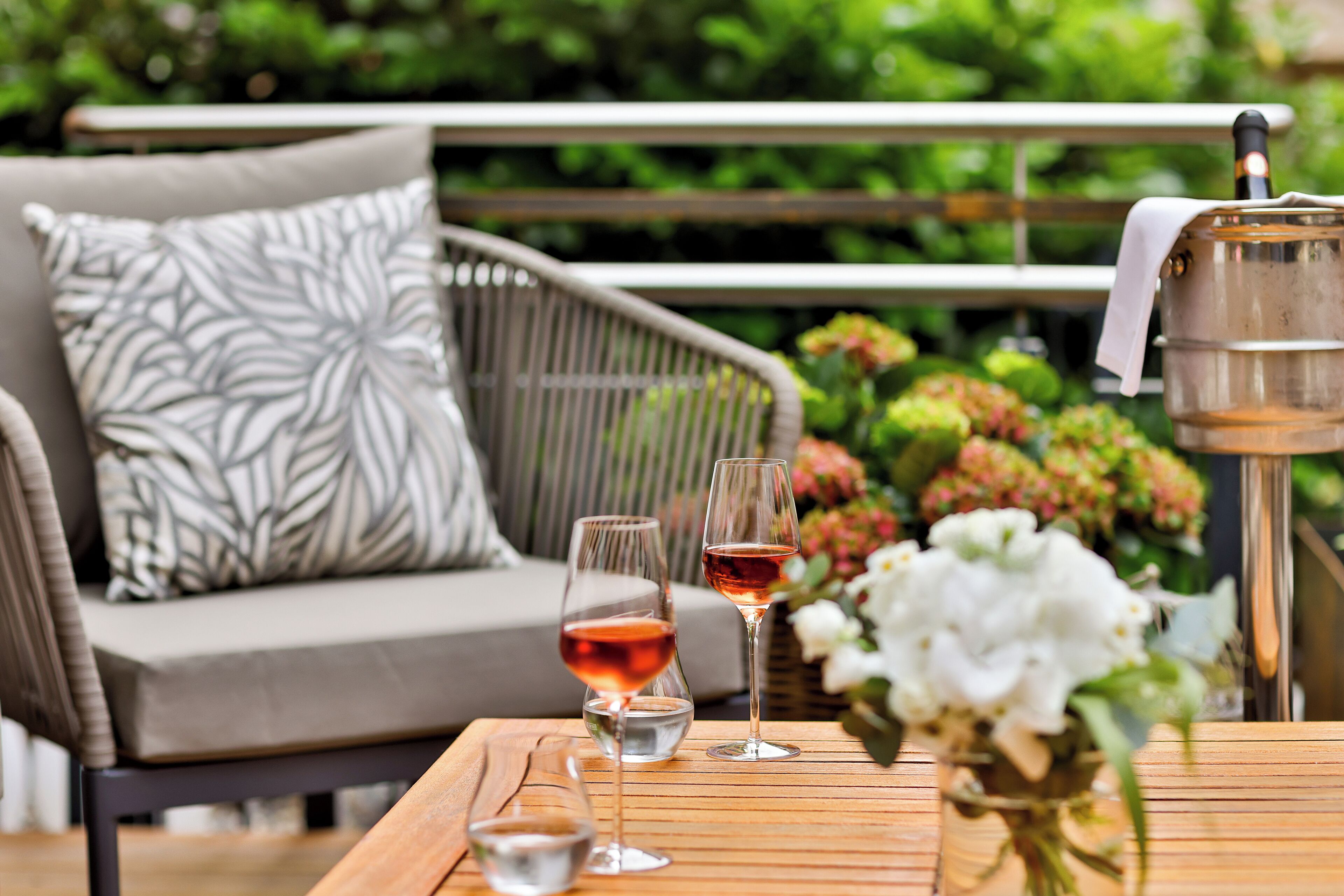 A wooden table with two wine glasses, a small bowl, and a metal container with a cloth on a patio with a wicker sofa and patterned cushions in the background.