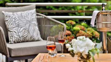 A wooden table with two wine glasses, a small bowl, and a metal container with a cloth on a patio with a wicker sofa and patterned cushions in the background.
