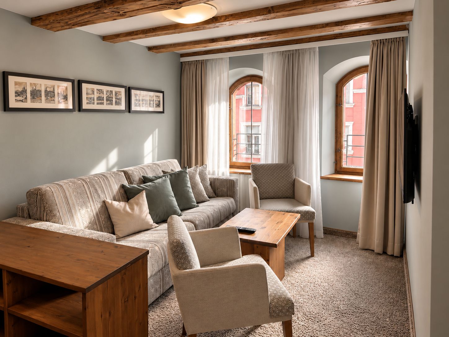 Ein gemütliches Wohnzimmer mit einer Couch, Sesseln, einem Couchtisch und einem Fernseher. An den Wänden hängen gerahmte Bilder und es gibt ein Fenster mit Vorhängen.