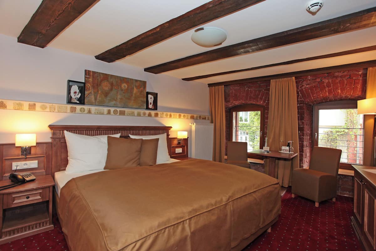 A hotel room with a large bed, a desk, a chair, a radiator, and a window with curtains.