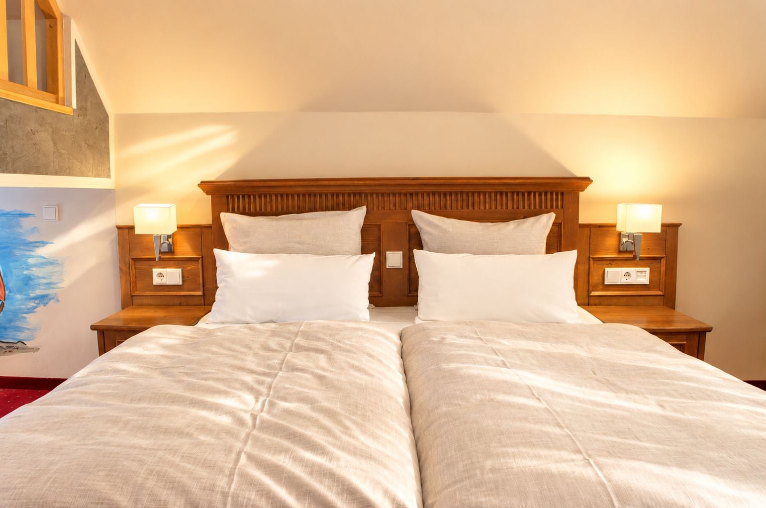 A double bed with two pillows, wooden headboard, bedside lamps, and a painting on the wall.