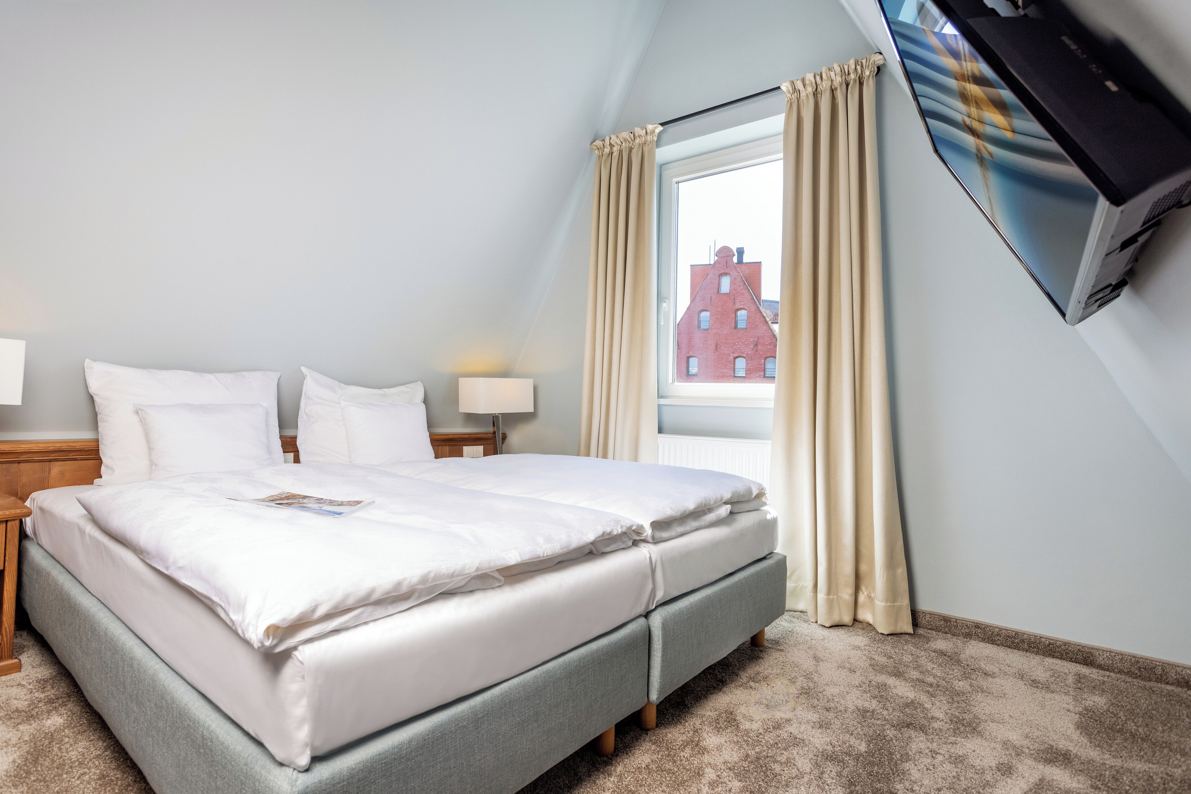 Une chambre d’hôtel avec un lit, un téléviseur fixé au mur, une fenêtre avec des rideaux et une vue sur un bâtiment à l’extérieur.