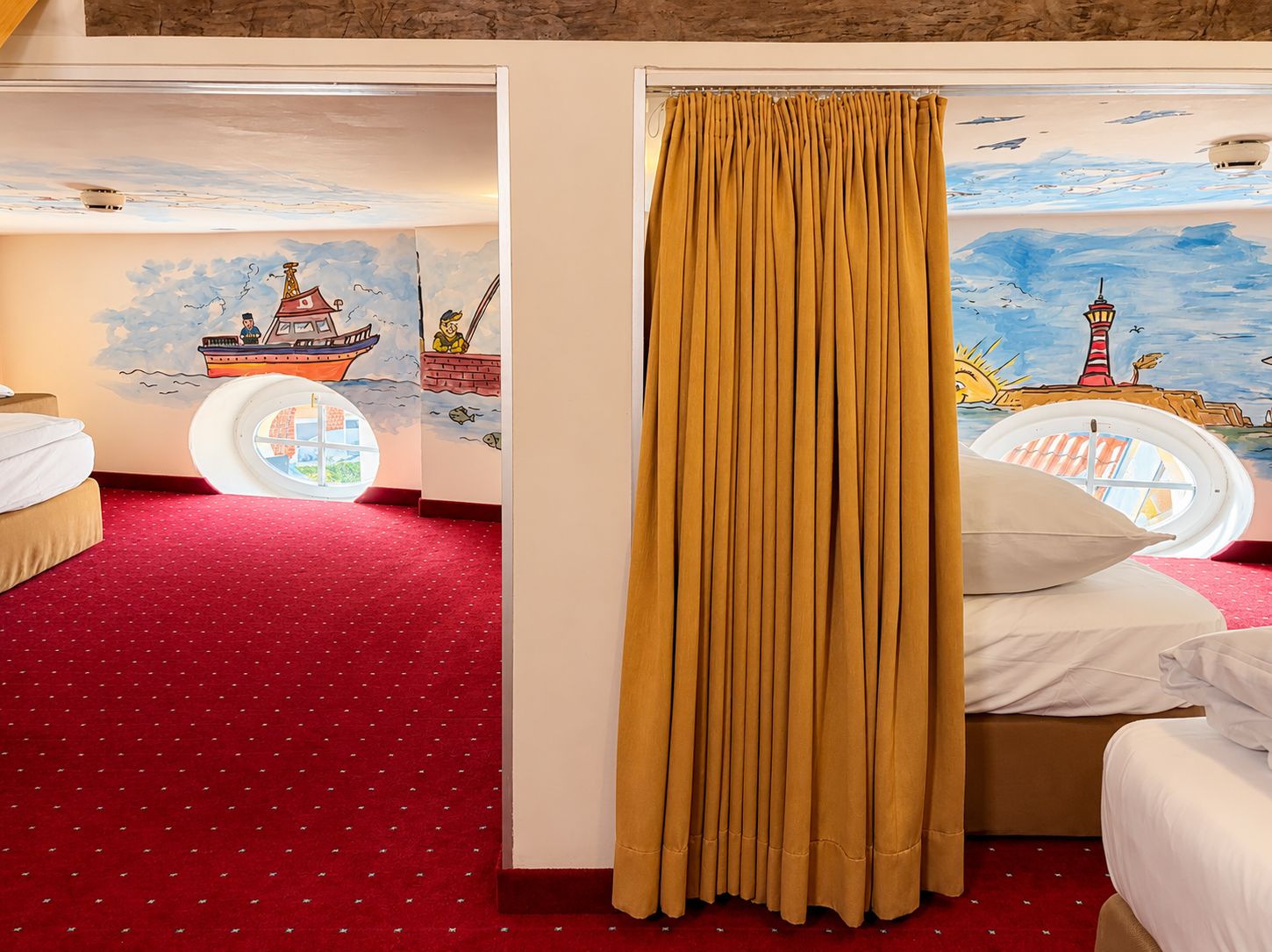 Habitación de hotel con alfombra roja, cama con ropa de cama blanca, ventana con vista a un faro y un mural que representa un barco y un faro.