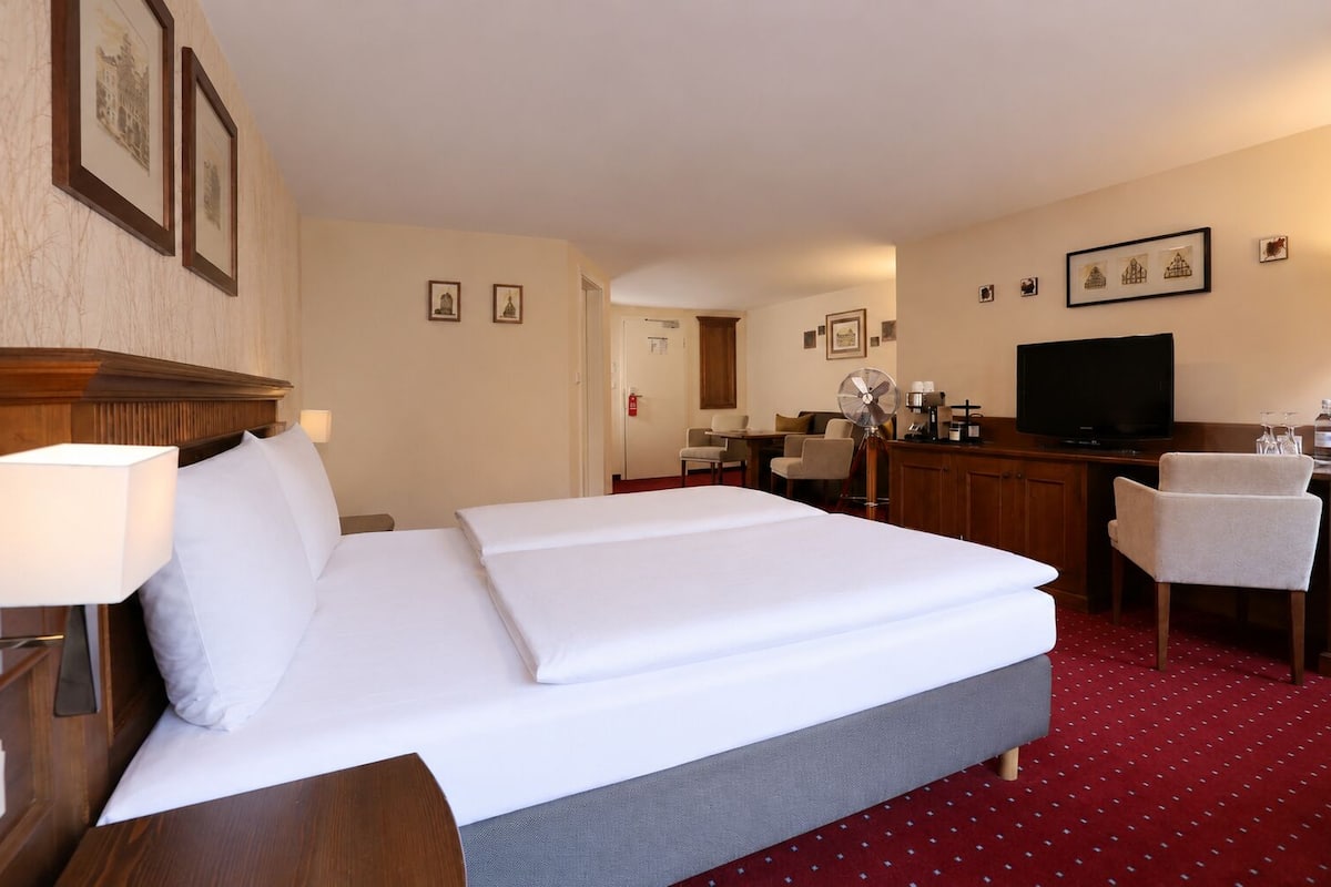 A hotel room with a double bed, a desk with a chair, a television, and a small kitchenette area.