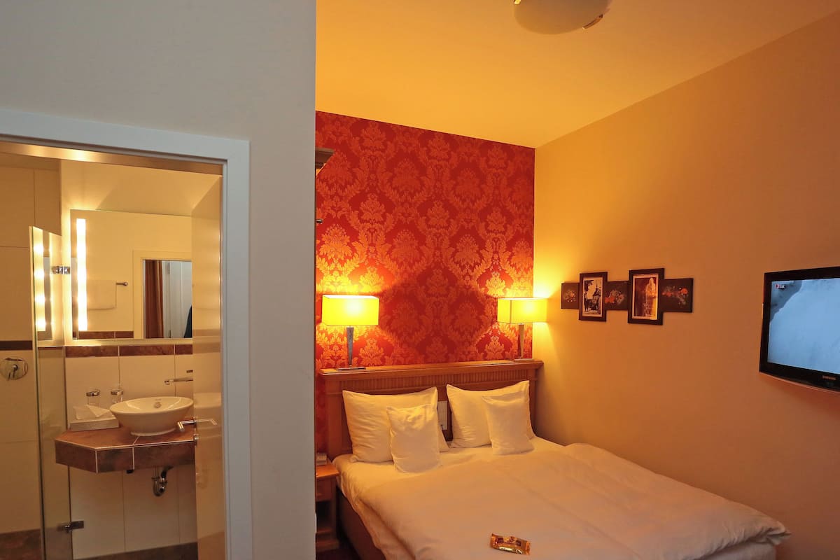 A hotel room with a bed, a bedside table, a TV, and a bathroom visible through an open door.