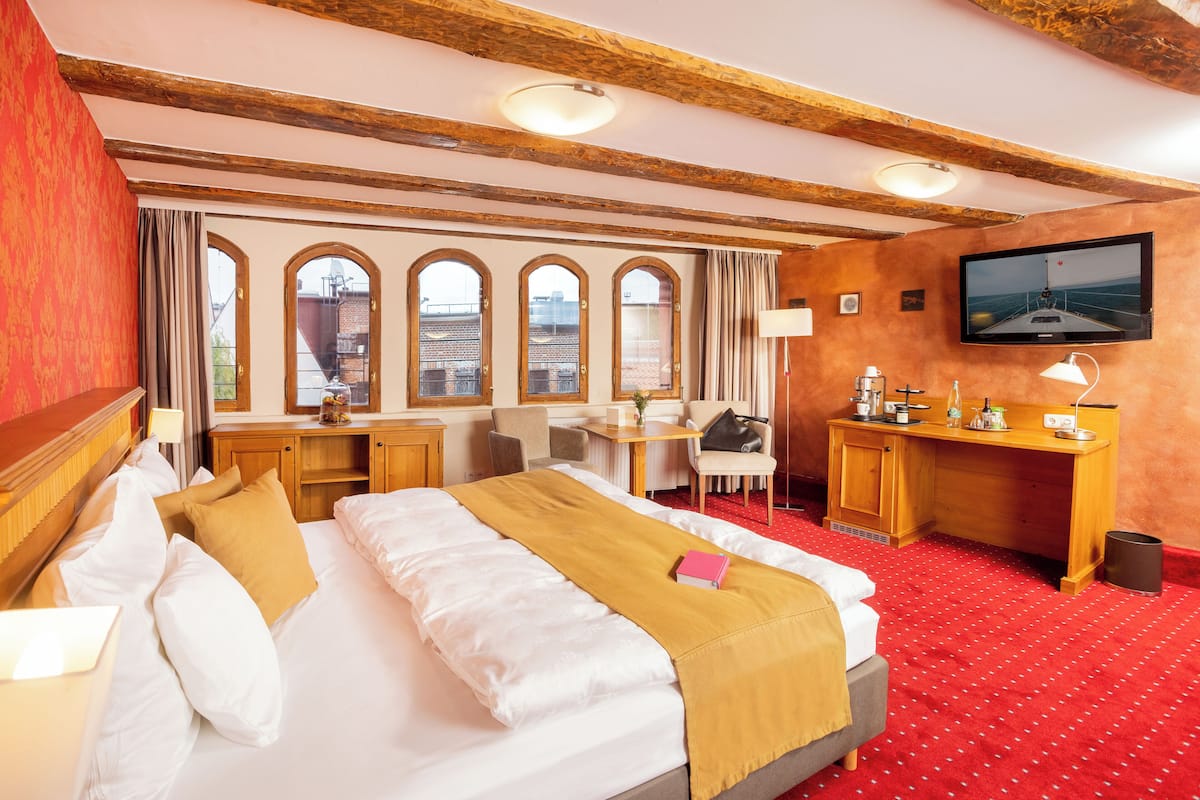 A hotel room with a large bed, a desk, a chair, a TV, and large windows with arched tops.