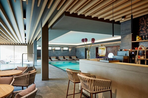 Un espace bar moderne avec une piscine en arrière-plan, des fauteuils confortables et un comptoir de bar où sont exposés divers articles.