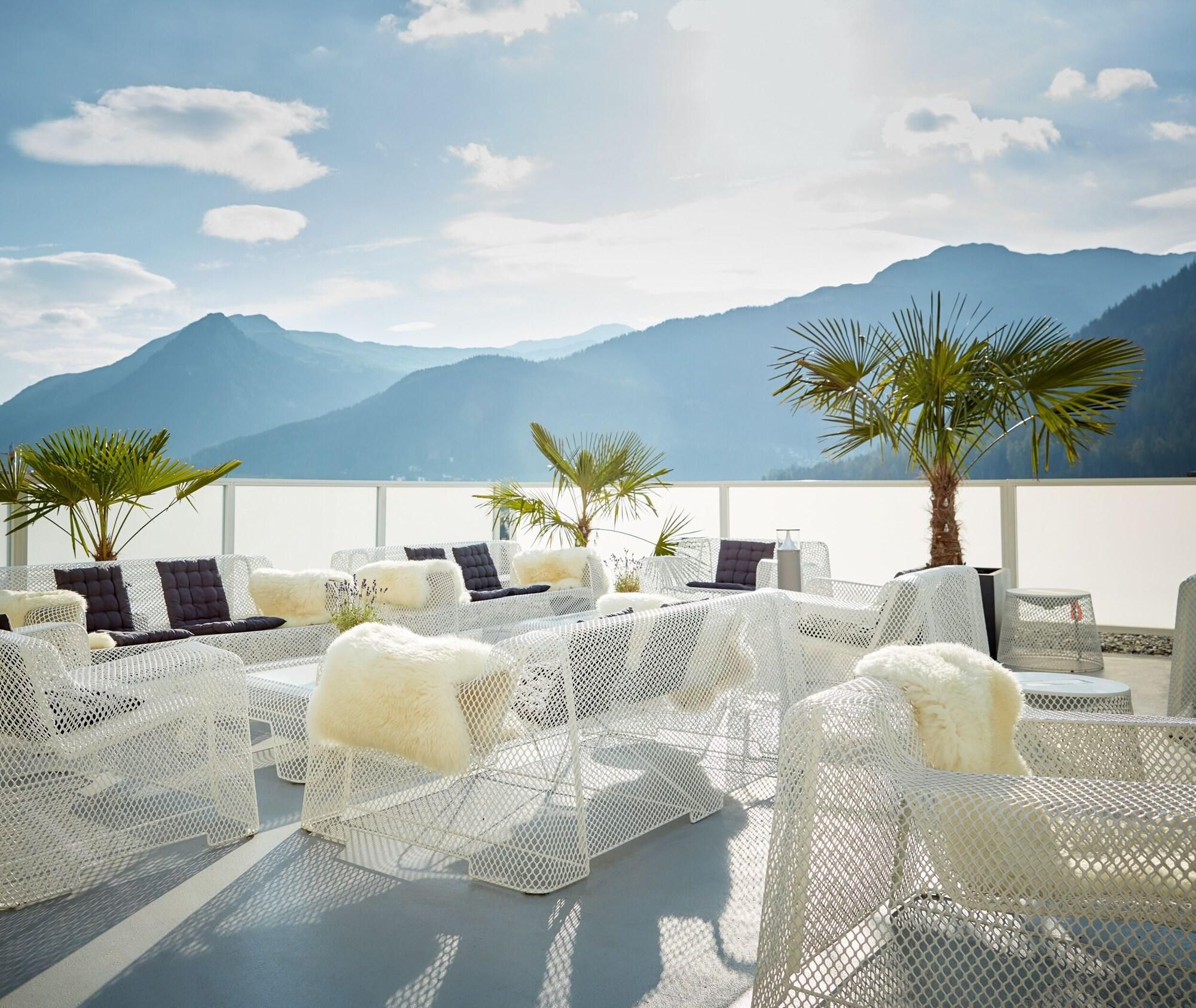 A rooftop lounge with white mesh chairs, some with fur throws, and a view of mountains in the background.