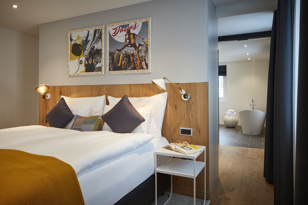 Ein Hotelzimmer mit Bett, Nachttischlampe, einem kleinen Tisch mit einem Buch und einem Poster an der Wand.