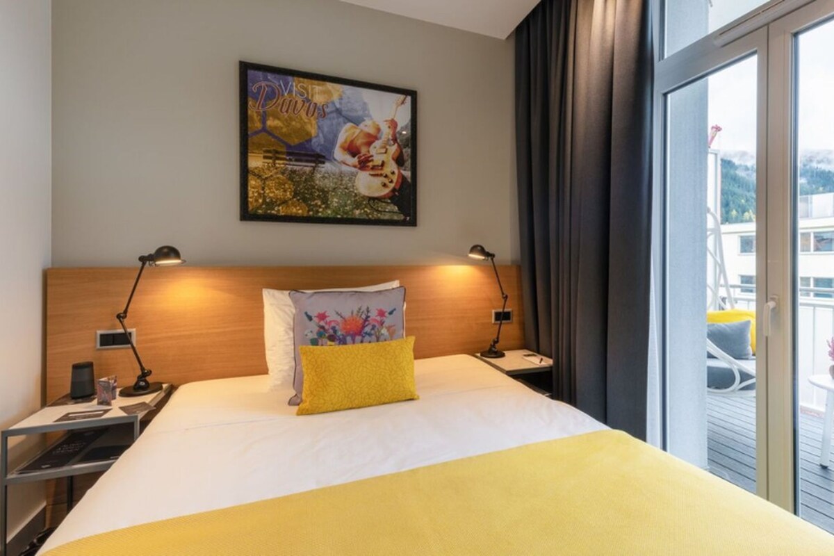 Ein Hotelzimmer mit einem Bett, zwei Nachttischen, einer Lampe, einem Gemälde und Blick auf einen Balkon.