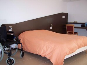 A single bed with an orange cover, a wheelchair, a desk, and a chair in a room with a brown headboard and white walls. - Hôtel Le Richevaux (Neufchateau)