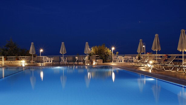A swimming pool with lounge chairs and umbrellas, surrounded by a fence and trees. - Asteris Hotel (Kefalonia)