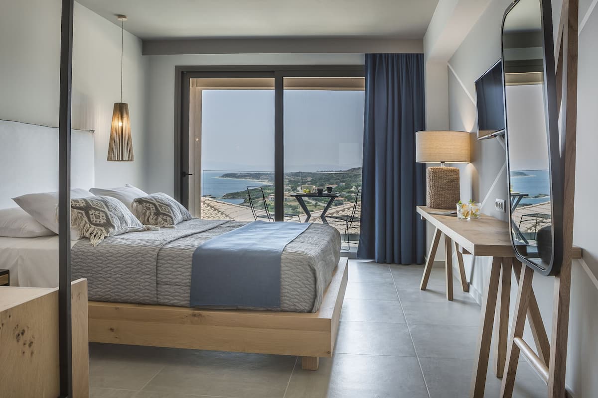 A modern hotel room with a large bed, a wooden desk, a TV, and a view of the sea.