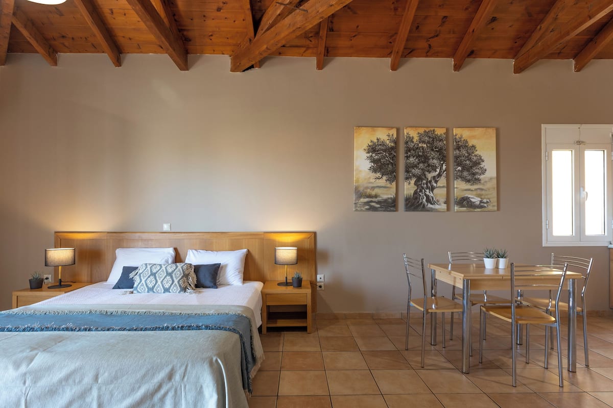 A bedroom with a wooden ceiling, a bed with a headboard, bedside tables with lamps, a dining table with chairs, and a window.