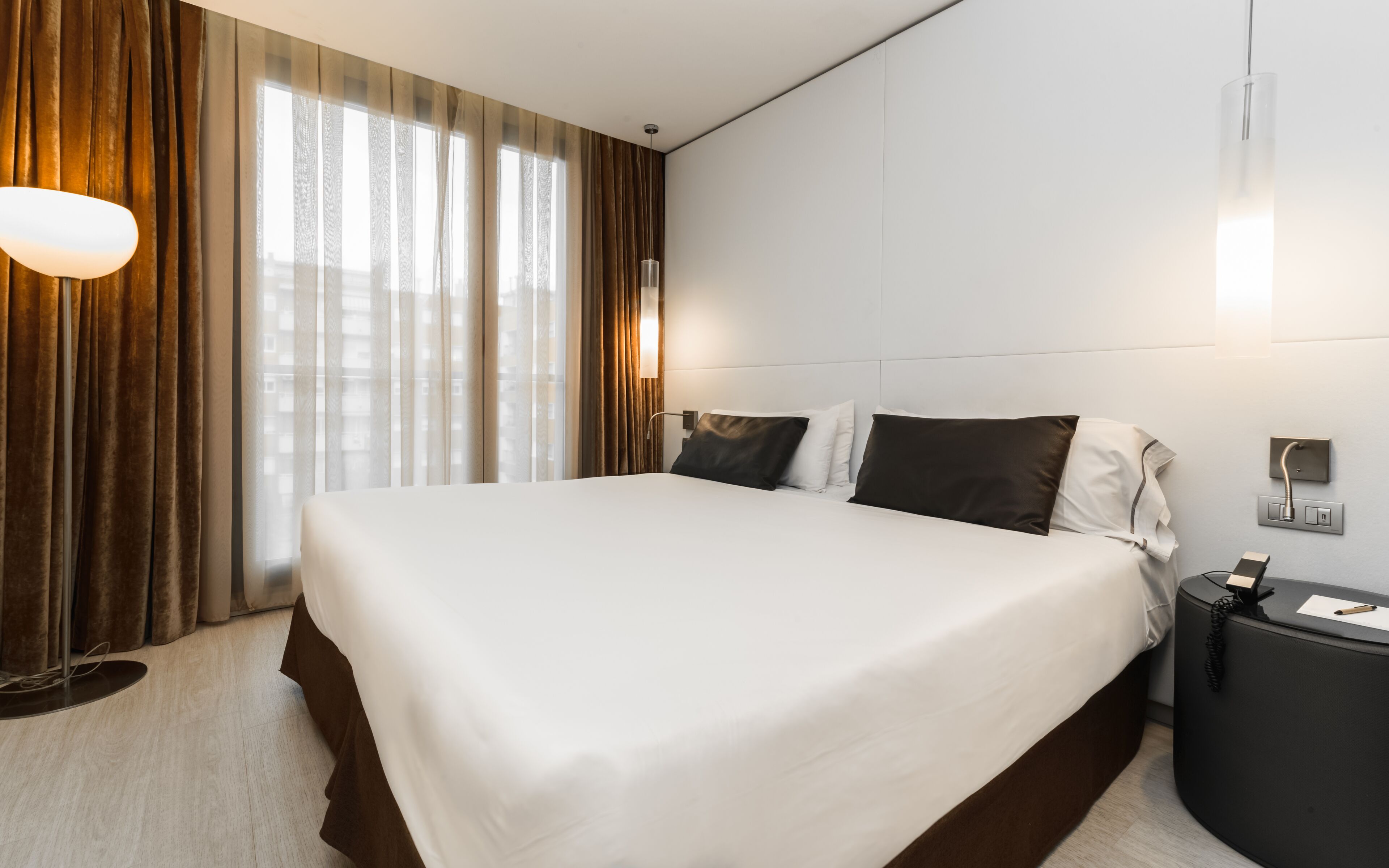 Ein Hotelzimmer mit einem großen Bett, einem Nachttisch, einer Lampe und Blick auf die Stadt durch leichte Vorhänge.