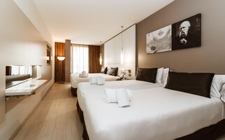 Quarto de hotel com duas camas, uma escrivaninha, um espelho e uma janela grande com cortinas.