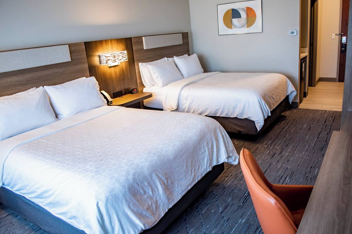 A hotel room with two beds, a desk, a chair, and a wall-mounted light fixture.