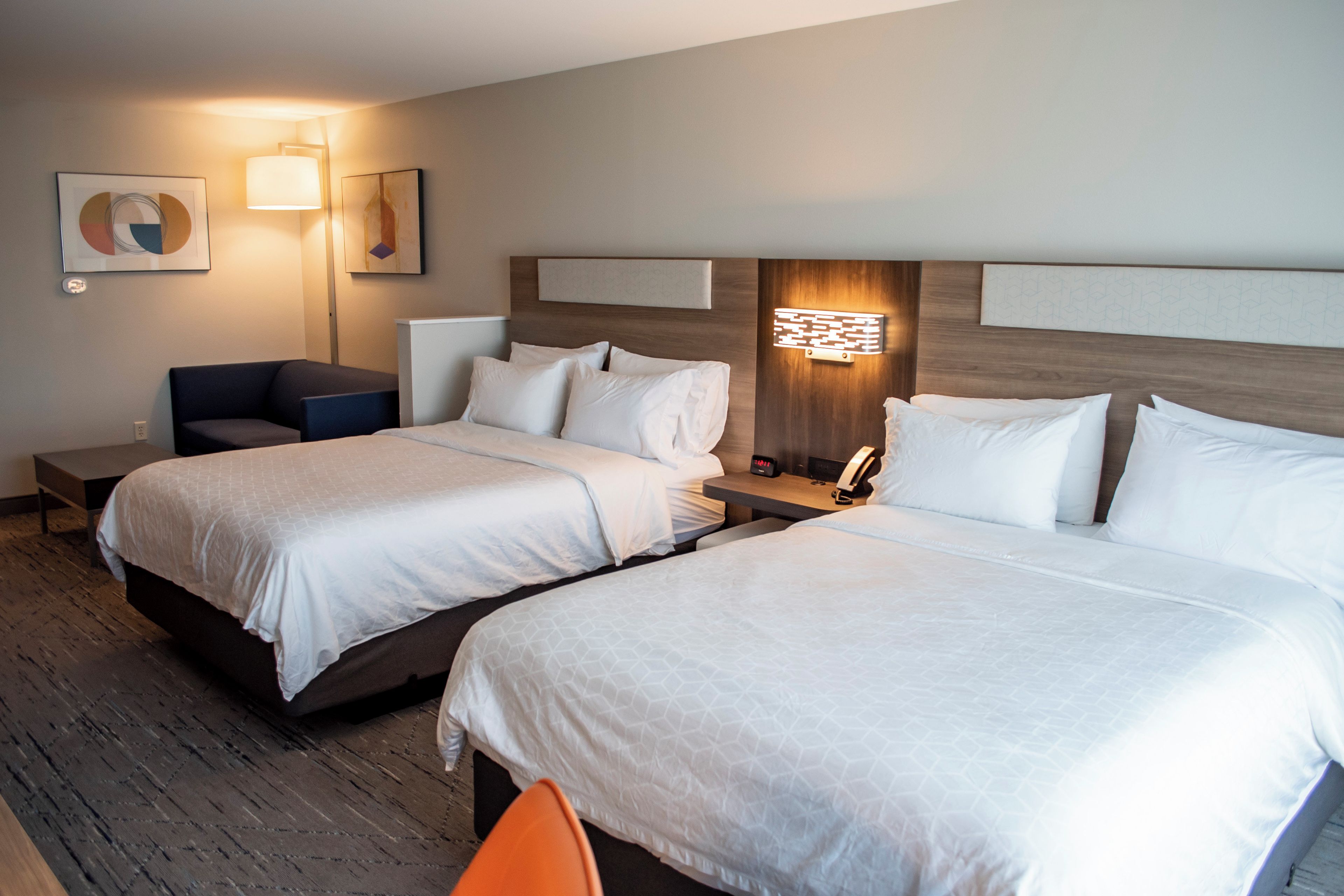 A hotel room with two beds, a desk, a chair, and a nightstand with a lamp.