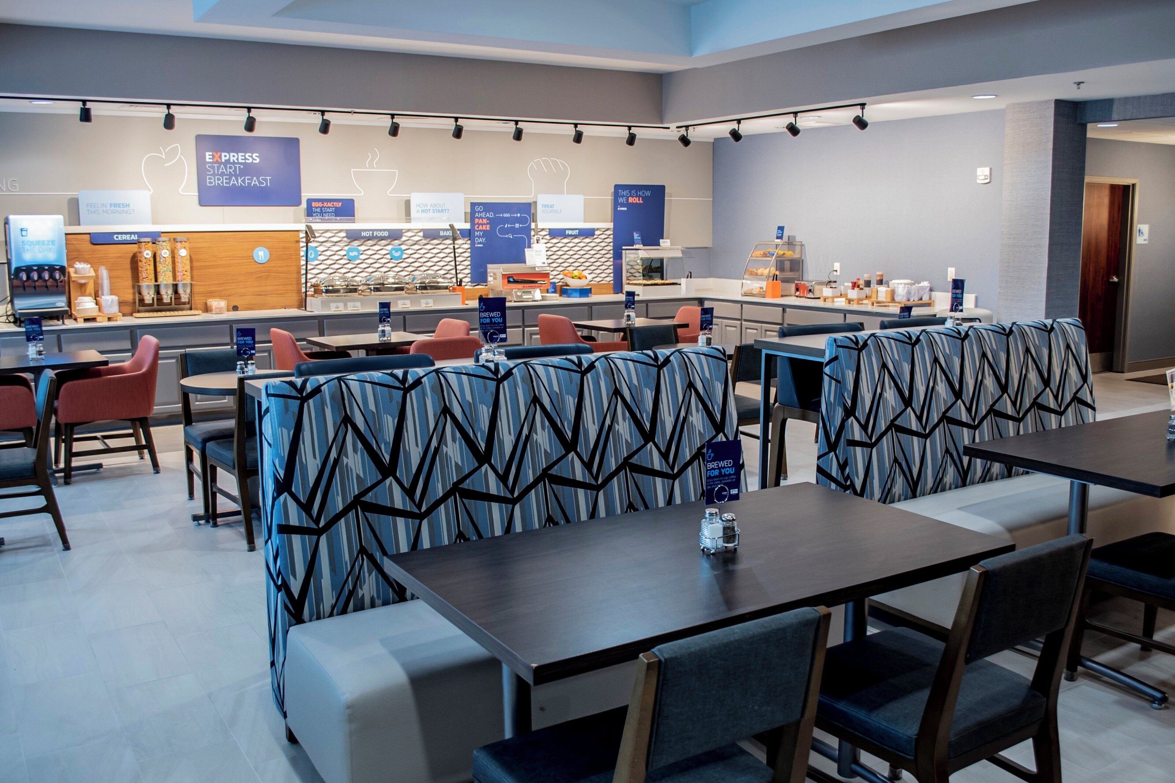 A modern hotel breakfast area with seating, a counter with food and beverage options, and a sign indicating breakfast availability.