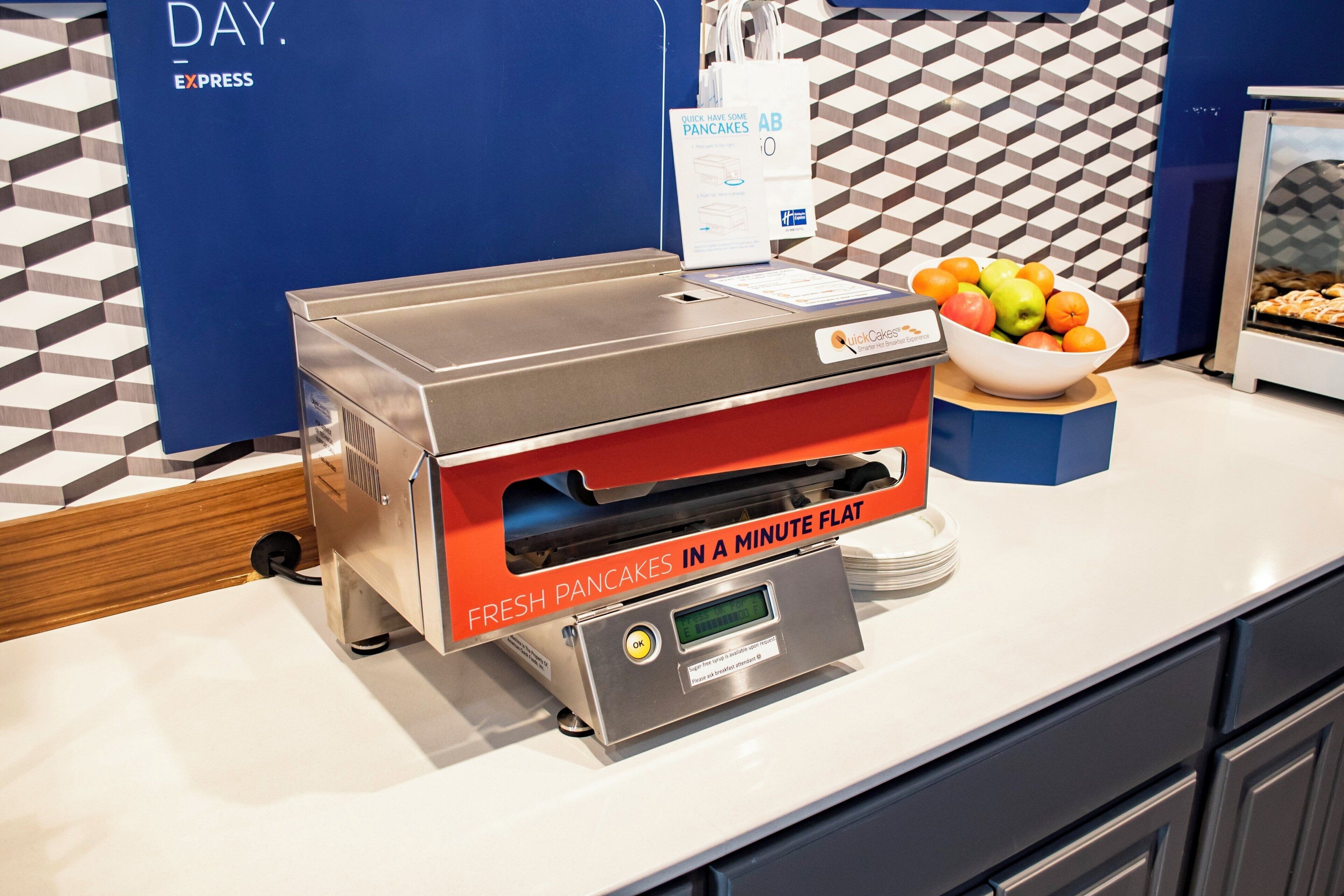 A breakfast station with a pancake maker, a bowl of fruit, and a sign indicating fresh pancakes in a minute flat.