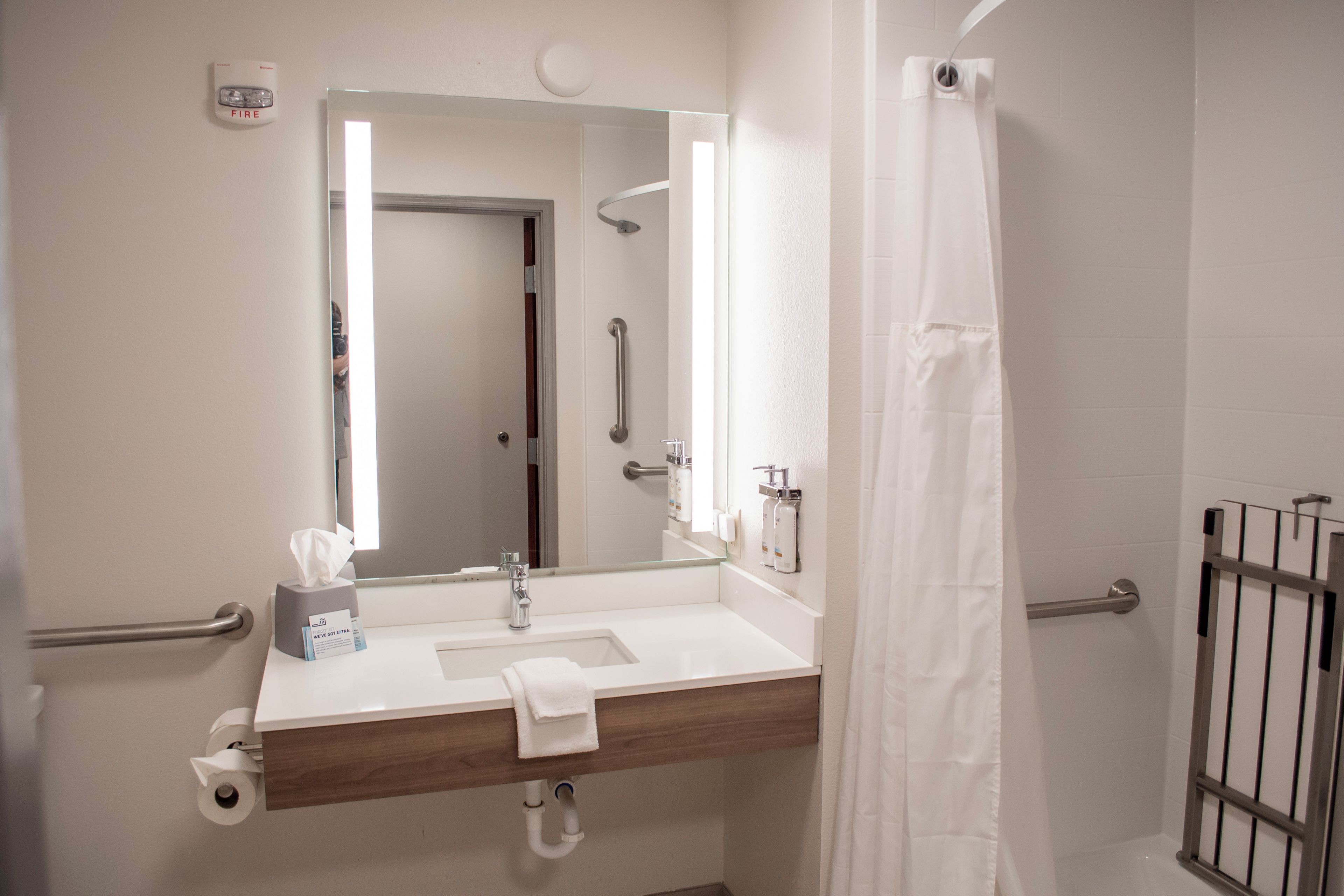 Un baño con lavamanos blanco, un espejo, una toalla, dispensador de papel higiénico y cortina de ducha.