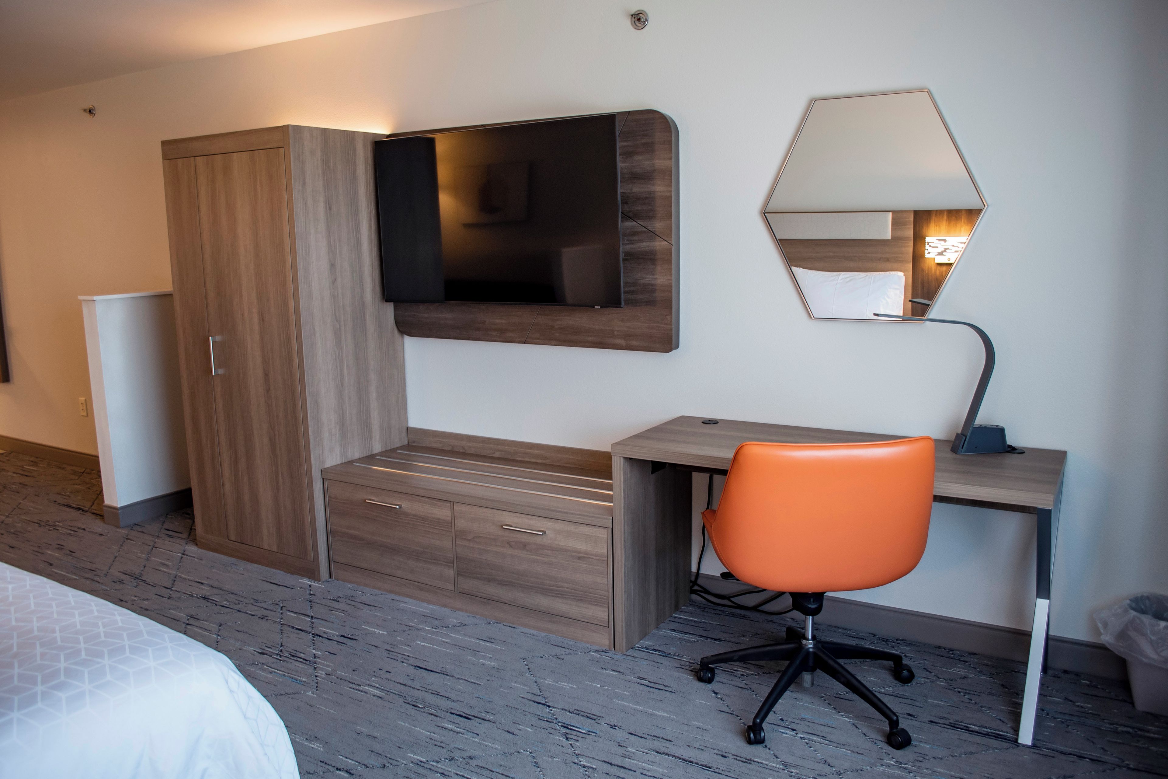 A hotel room with a bed, a desk with an orange chair, a wardrobe, a flat-screen TV, and a mirror.