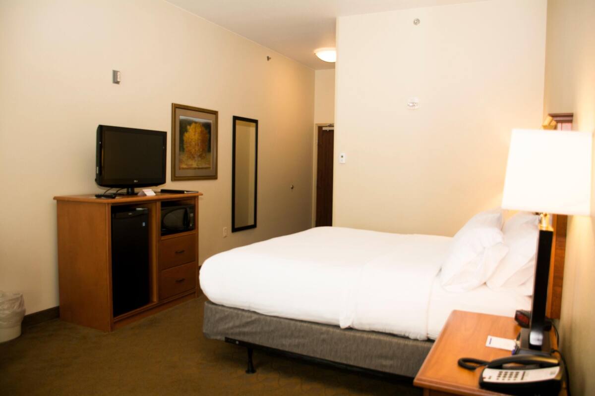A hotel room with a bed, a desk with a lamp, a television, a mirror, and a door.