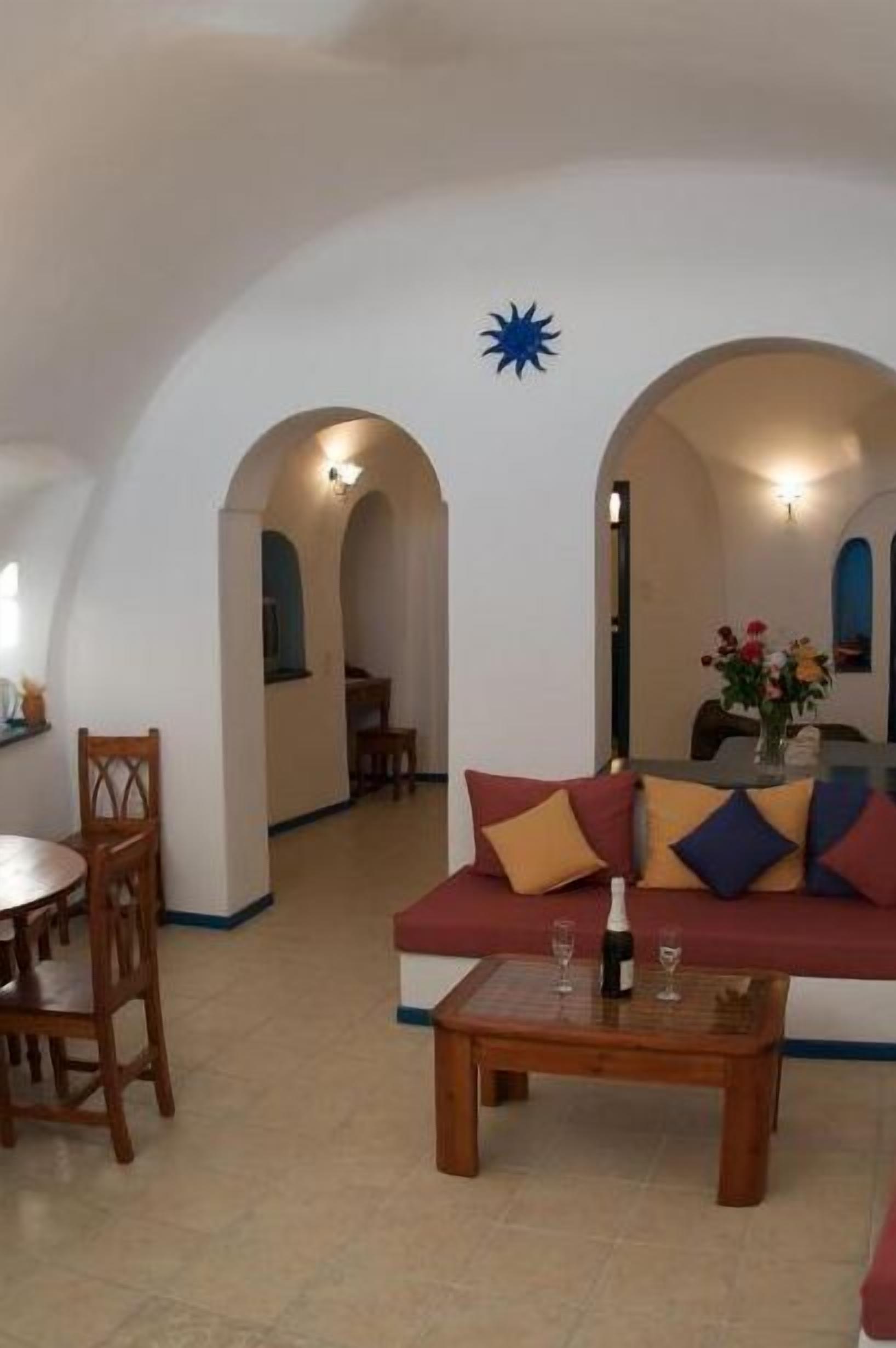 Uma sala de estar acolhedora com sofá, mesa de centro e mesa de jantar com cadeiras. Há portas em arco que levam a outros cômodos.