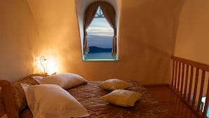 A bedroom with a bed, pillows, a nightstand, a window with curtains, and a wooden railing. - Vallas Apartments & Villas (Santorini)