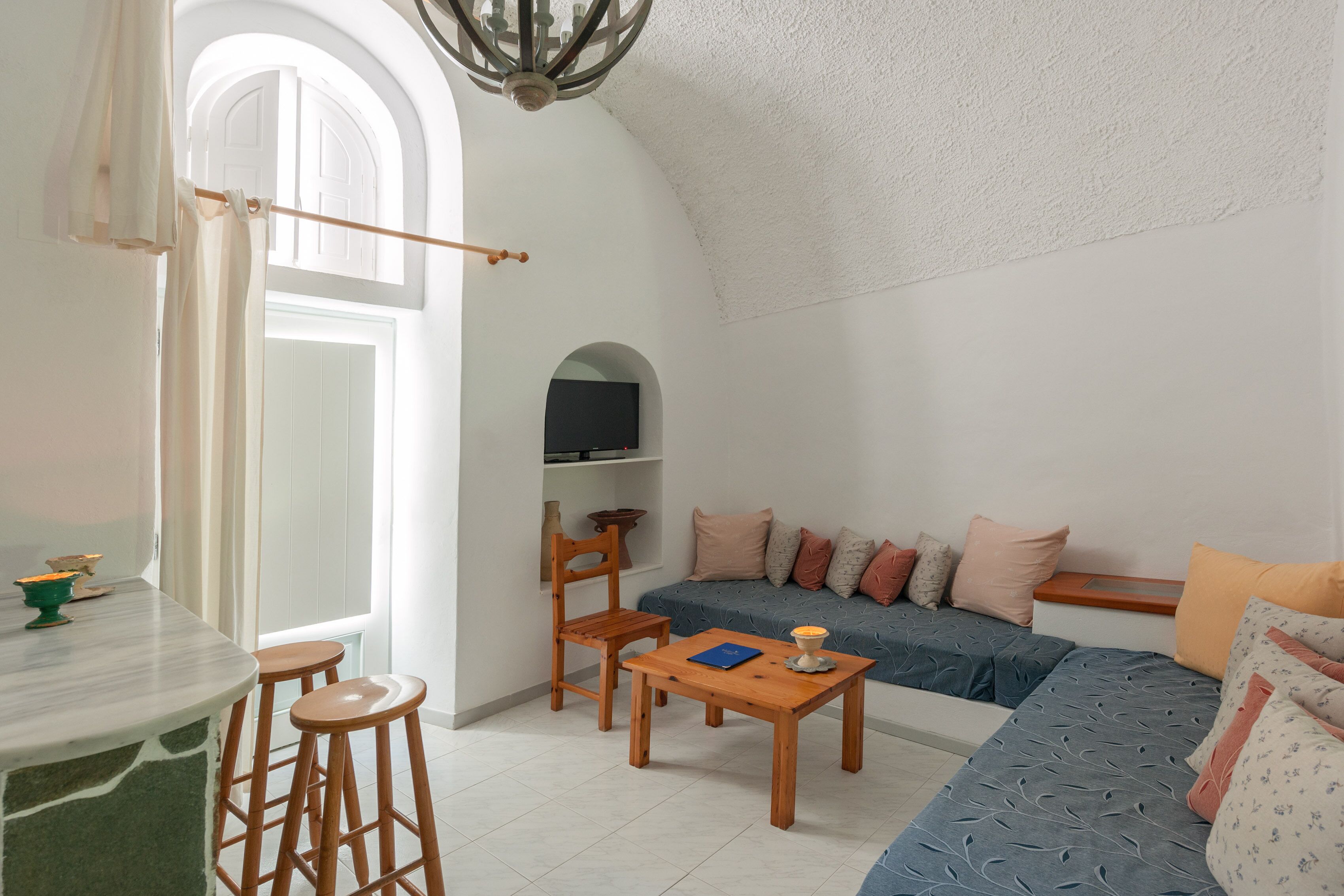 A cozy living area with a sofa, a wooden table, and a chair. There is a window with curtains, a small table with a candle, and a shelf with decorative items.